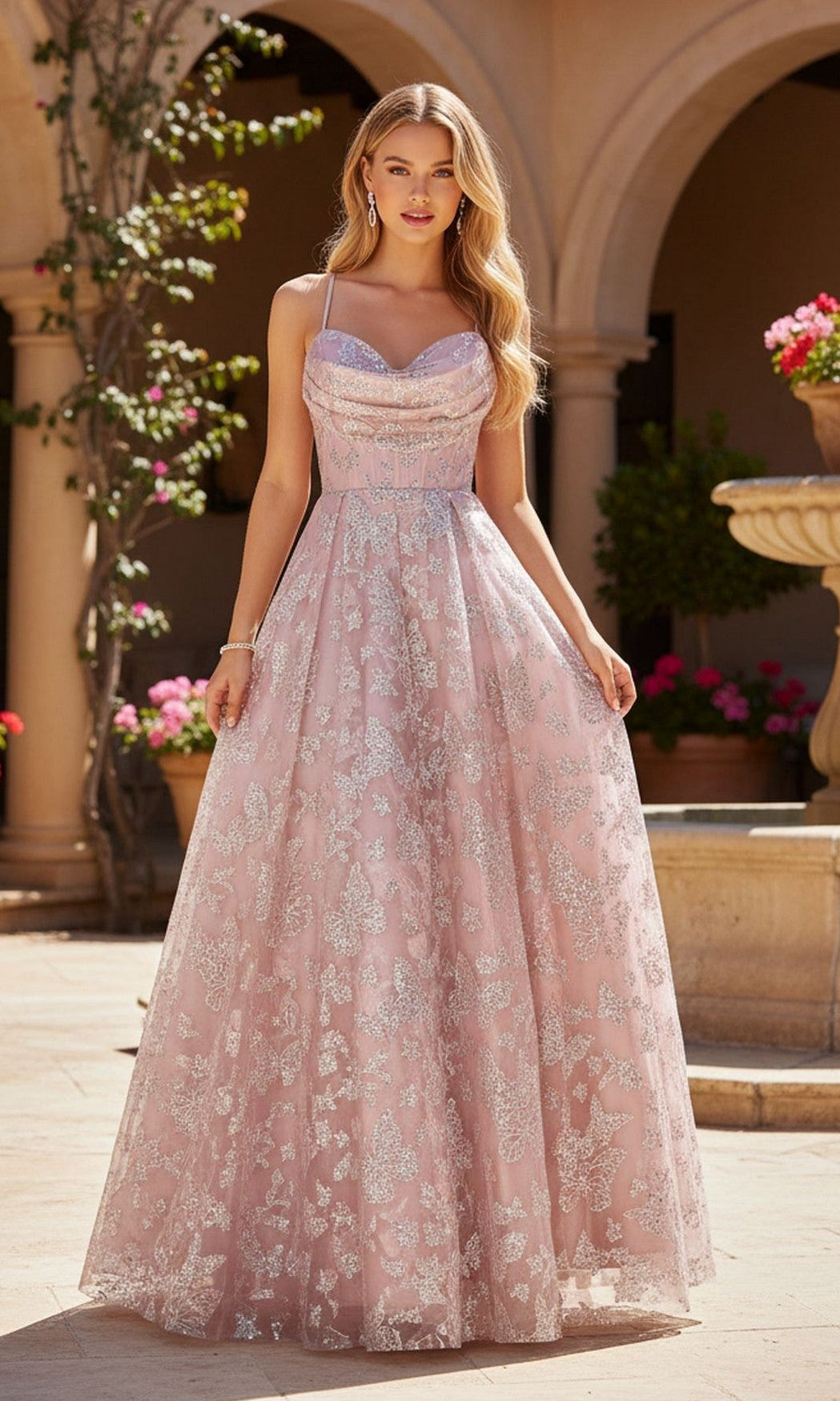 A woman in the Blondie Nites 6007BN long prom dress in rose, featuring a corset bodice and silver floral accents, stands outdoors by a stone fountain and potted flowers.